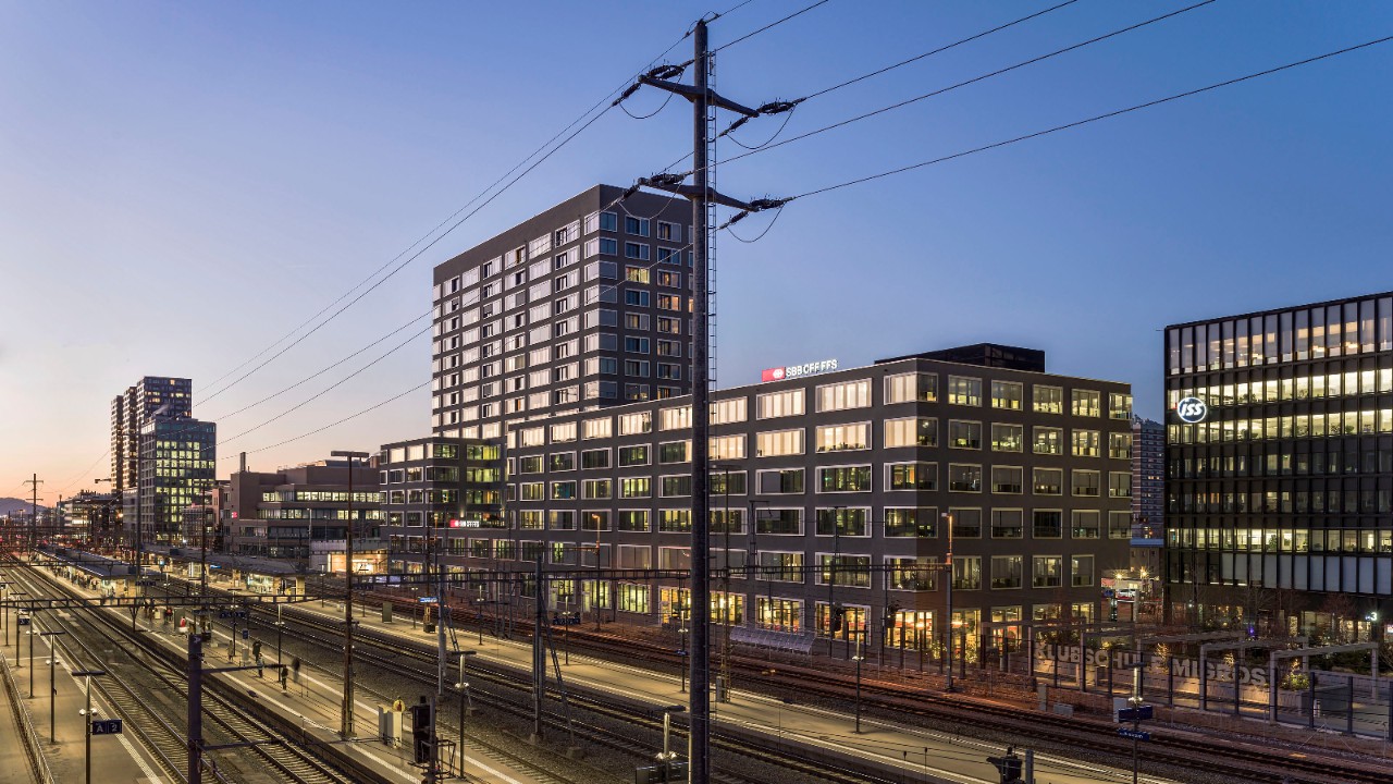 Das Bild zeigt mehrere beleuchtete Bürogebäude neben einem Bahnhof in der Abendstimmung.