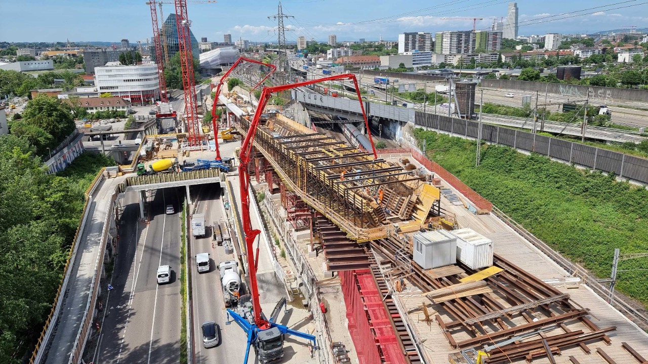 Das Foto zeigt den Bau der Donnerbaumbrücke in Muttenz. Zwei rote Kräne unterstützen von der Strasse aus den Bau des Gerüsts für die Betonarbeiten. Rechts davon verlaufen die Bahnlinien.