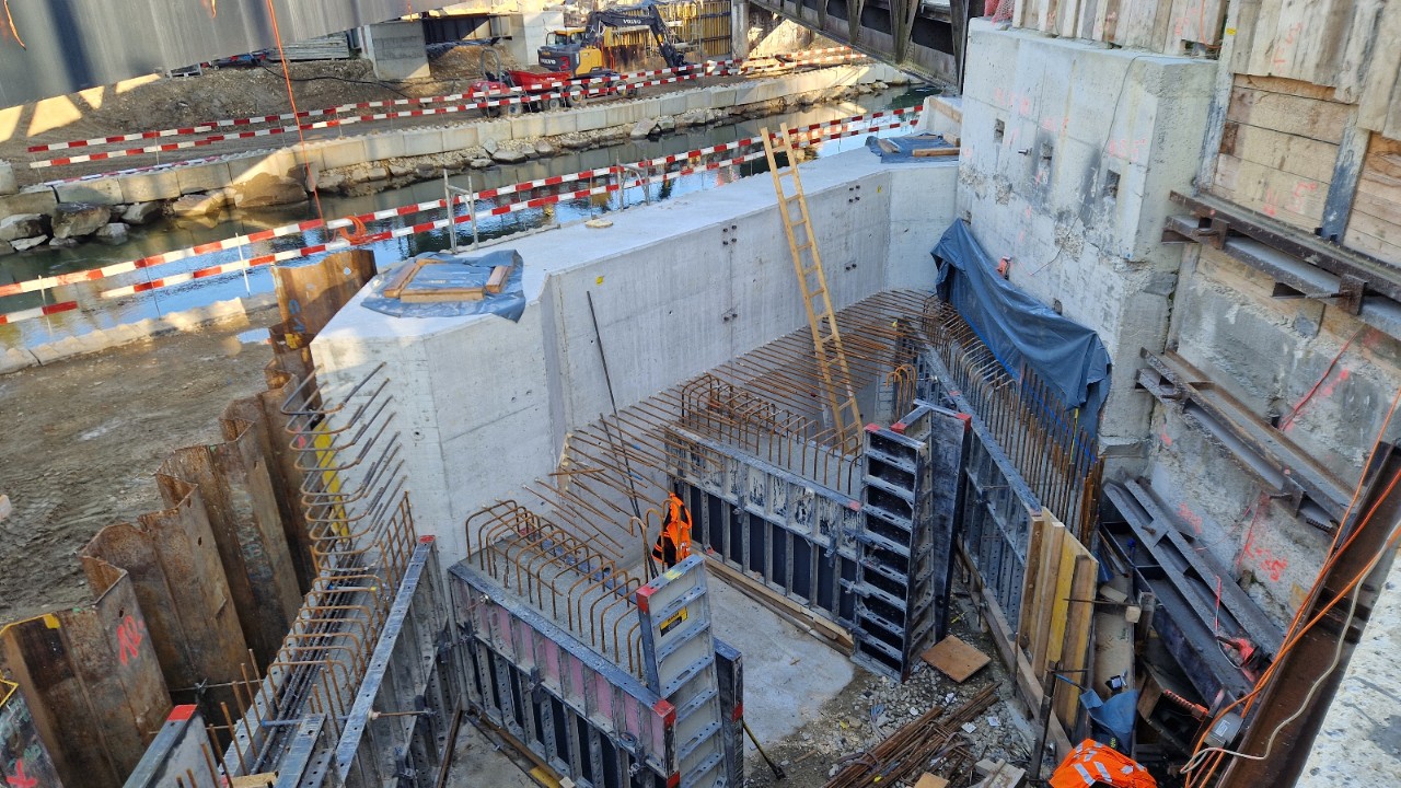 Das Bild zeigt eine Baustelle mit Betonmauern, Verschalungen und Armierungseisen. Weiter hinten im Bild ist ein Fluss und weitere Betonarbeiten zu sehen. 