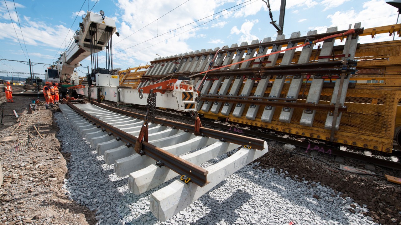 Das Bild zeigt ein Beispiel einer Fahrbahnerneuerung: Mit einer grossen Gleisbaumaschine werden Gleise samt Schwellen auf einem Schotterbett platziert.